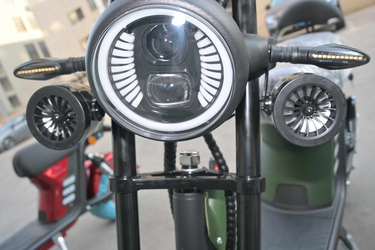 Vista frontal aproximada da scooter, destacando o farol redondo de LED com luz diurna integrada e os dois alto-falantes Bluetooth pretos instalados nas laterais do garfo dianteiro.