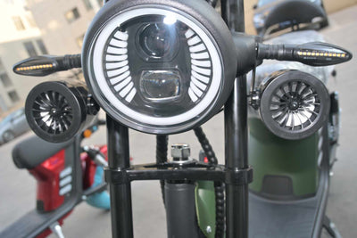 Vista frontal aproximada da scooter, destacando o farol redondo de LED com luz diurna integrada e os dois alto-falantes Bluetooth pretos instalados nas laterais do garfo dianteiro.