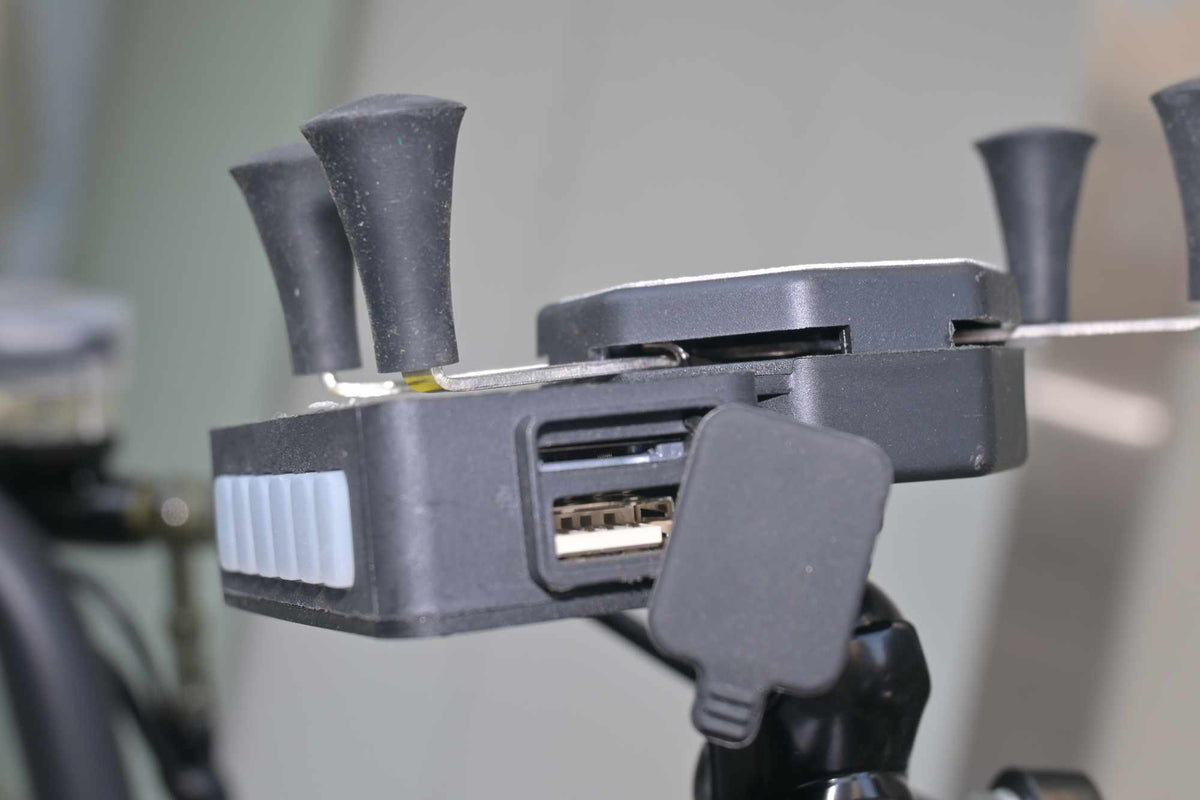 Vista lateral em close do suporte para celular, mostrando a tampa de borracha aberta que revela uma porta USB integrada para carregamento de dispositivos móveis.