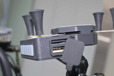 Vista lateral em close do suporte para celular, mostrando a tampa de borracha aberta que revela uma porta USB integrada para carregamento de dispositivos móveis.